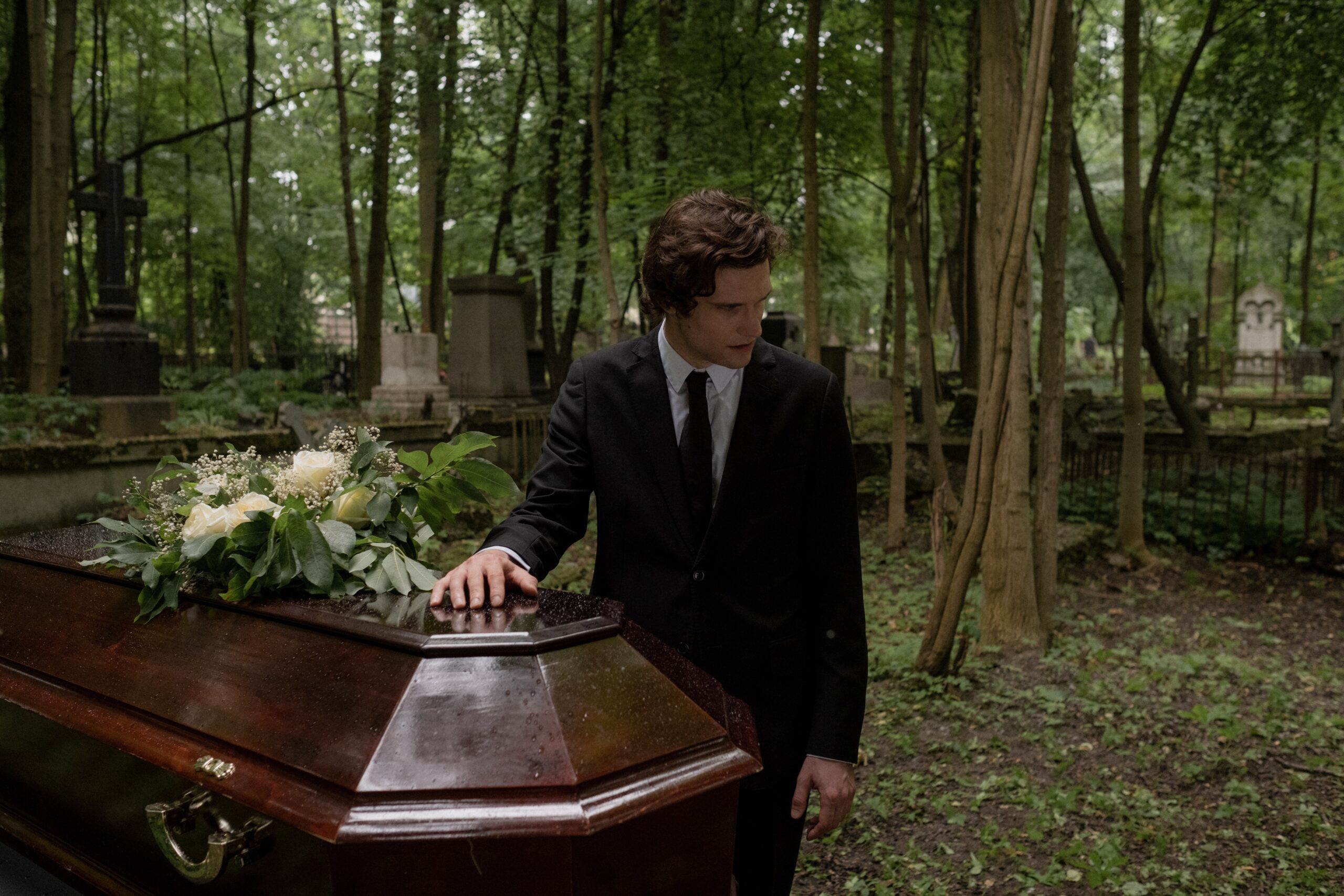 Mężczyzna w czarnym garniuturze stoi na cmentazu trzymając dłoń na trumnie